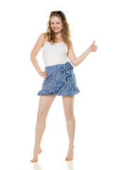 Young blonde woman giving thumbs up, wearing white top and blue floral skirt. Energetic, positive full-body pose with bright smile on neutral background