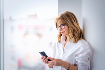 Blond haired mature woman holding cell phone in her hand and standing at wall by the window