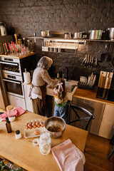 A light-skinned gray-haired grandmother in her 60s washes the dishes at a sink in a well-equipped decorated with hanging string lights kitchen, with her school-aged granddaughter in a colorful sweater