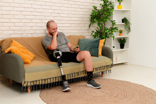Man amputee with bionic prosthesis leg disability spending time at home, sitting on the sofa watching sports movie on smartphone, concentrated expression