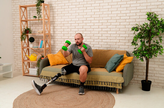 Man amputee with bionic prosthesis leg disability spending time at home, sitting on the sofa doing sports fitness exercise with dumbbells.