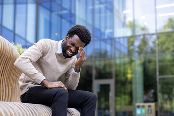 A black man is experiencing a painful moment while sitting on a modern bench outside a building....