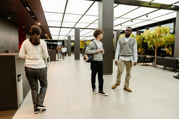 Two interns in their 20s, a White man in glasses and a cardigan with a backpack, and a Black man in a sweater vest with a laptop walk and talk in a modern office lobby with trees and other workers.