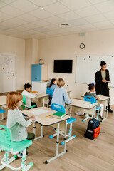 A Black female teacher aged 35, dressed in a black business suit, talking to her first-grade students while standing next to the whiteboard in her classroom, as one of the boys gets up from his desk