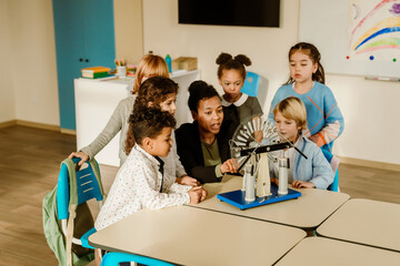 White, Black, and Latino children gather around a table in a classroom with white walls to conduct...