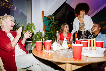 A woman standing next to a multiethnic group of three friends consisting of two women and one man sitting at a table with a cake and red plastic cups during a birthday party and laughing