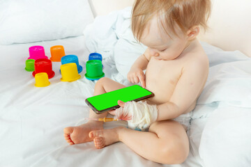 child in hospital sit in bed with mobile phone smiling. baby plays on the phone with intravenous infusion set to drop. concept of courage and joie de vivre. mockup with a green screen