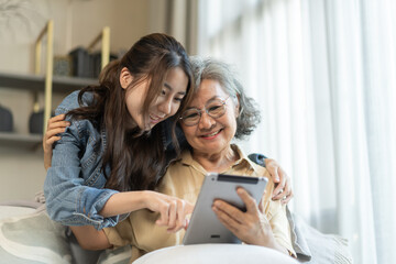 Asian senior mother and daughter smiling and talking using tablet browsing social media, shopping online, watching movies or series, enjoying entertainment digital connection and bonding at home