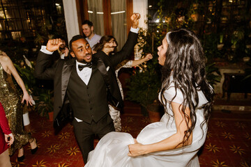 A Black groom in his 20s in a black suit dances near a Black bride in her 20s in a white dress, in a wedding conservatory with plants at night, with other multiethnic guests dancing in the background.
