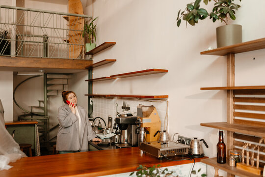 In a modern cafe, a White businesswoman in her 20s with red hair and a grey blazer stands by a kitchen area behind a counter talking on a phone near coffee equipment, shelves, plants, and stairs.