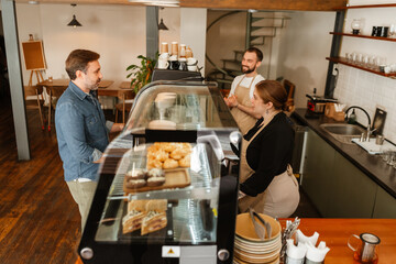 Three people, two men, and one woman, interact while working in a cafe with a display case with pastries and baked goods. An adult White woman with red hair in a beige vest stands behind the counter.