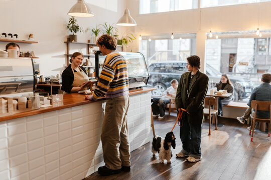 A young, short White businesswoman in her 20s, wearing an oversized suit, with her small dog on a leash, standing in a cozy coffee shop and waiting to make the order