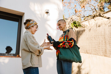 Outside a house, a smiling White woman in her 50s wearing a bandana and a beige shirt accepts a wine bottle from a smiling White woman in her late 40s with a shaved head wearing a colorful sweater.