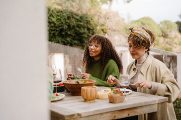 A hipster Latina woman in her early 30s, with glasses and a nose piercing, sitting at a wooden table on the porch of a house, chatting with her friends who sit around her, as they're having dinner