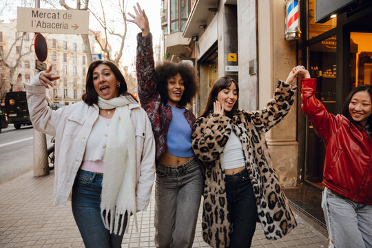 On a city sidewalk, four smiling multiethnic stylish women in their late 20s, including Black, White, Asian, and Hispanic, walk pompously, throwing hands in the air, some holding hands, during daytime