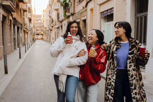 A young, short Asian woman aged 25, laughing cheerfully with her two young female friends as they walk along a city street, dressed in stylish outfits, holding bottles of soda