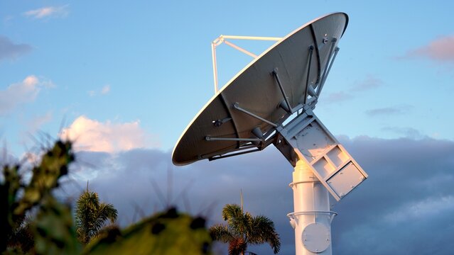 Large parabolic aerospace radar antenna pointing skyward, capturing signals from the cloudy atmosphere during a vibrant sunset, framed by palm trees in the foreground