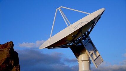 Large parabolic aerospace radar antenna directing toward a clear blue sky with fluffy clouds,...