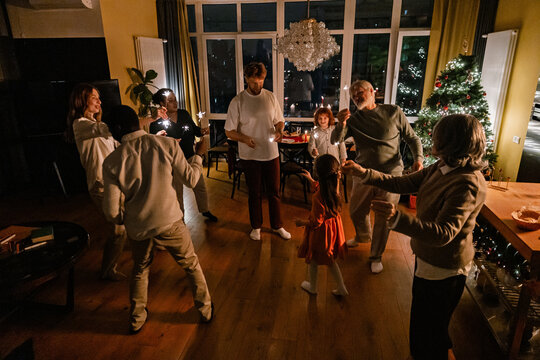 A multinational group of people of different ages, including children and elders, celebrating Christmas by dancing and playing with sparklers in a cozy living room with a decorated Christmas tree.
