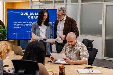 A White elder male office worker with gray hair and a beard, his Asian young female colleague, and...