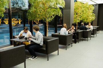 In a business center cafe, diverse workers sit in pairs on black sofas, having lunch, with small decorative trees between them. Concept of a lunch break, shared meals, and colleague interaction.
