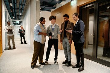A diverse group of adult men and women, including Indian, Black, White, and Middle Eastern coworkers, discuss in a modern office hallway with tablets and notebooks.