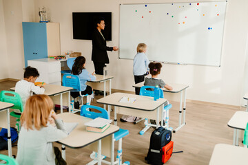 Obraz premium During a lesson in a classroom, a young Black teacher and a White boy stand at the whiteboard, while other children in the class sit at their desks. Concept of school activities, a study process.