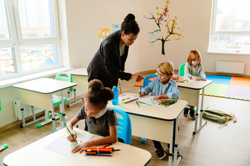 Obraz premium Black female teacher assisting diverse school-aged children drawing at desks in a brightly lit classroom, with boys and girls focused on their creative activity.