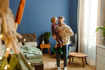 In the bedroom between the bed and the window, a White father in his 30s holds his school-age son in his arms and they both smile. The concept of fatherhood, childhood, weekend.
