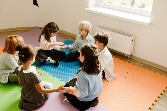 Children play the Stella Ella Ola clapping game while sitting in a circle on a colorful school mat, enjoying their time together. A concept of childhood entertainment and fun.