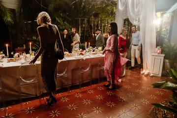 At an elegant outdoor venue, guests in stylish attire gather around a set dining table with candles and floral arrangements. Concept of a festive and intimate atmosphere, a celebratory event.