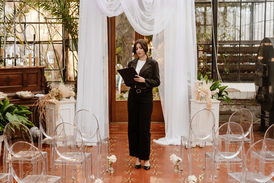 A White young female wedding planner standing on a red-tiled floor in a decorated venue aisle with transparent chairs and white drapery, reviewing notes on a clipboard before the ceremony begins.
