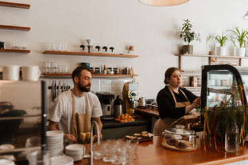 A White man and woman wearing aprons work behind the counter of a modern cafe. The man prepares food while the woman arranges items in a glass display case filled with desserts.