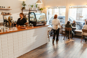 A White young female cafe owner stands behind the counter beside a pastry display while a White young female worker with red hair serves a group of seated customers inside a bright modern cafe.