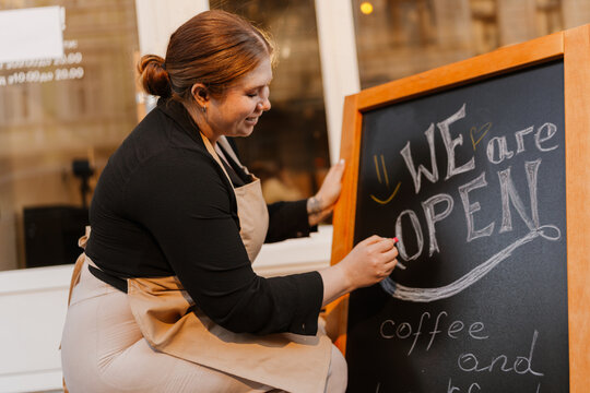 A white adult woman in an apron writes "We are open" on a chalkboard sign at a coffee shop entrance. She is smiling while preparing the front of the business for opening indoors.