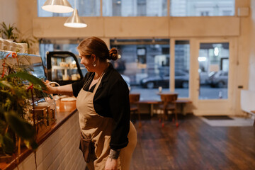 A young female cafe owner prepares the counter for closing time inside a warmly lit coffee shop with wooden flooring, modern hanging lamps, and large windows revealing the evening city street.