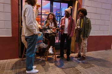 A multinational group of young friends standing and sitting outside a bar in the evening, drinking and chatting casually with warm outfits, one holding a small dachshund dog on her lap.