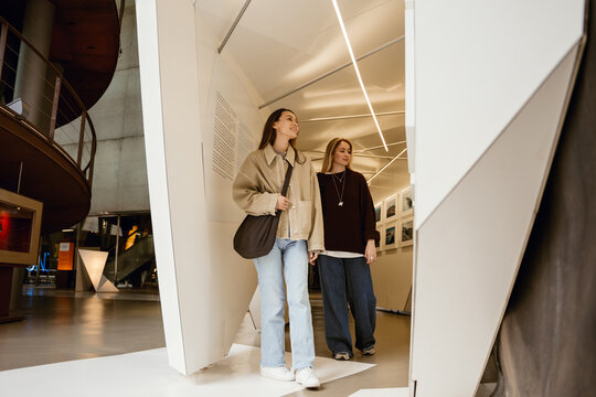 Two young White women stroll through a modern museum, looking at an exhibition with text and photographs on the walls. Concept of a cultural recreation, learning something new, meeting a friend.