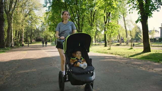 A young sporty woman runs in the park with her son in the running board. Mom is happy to do sports in the park with her newborn baby on a sunny day. Concept of power, fitness, family