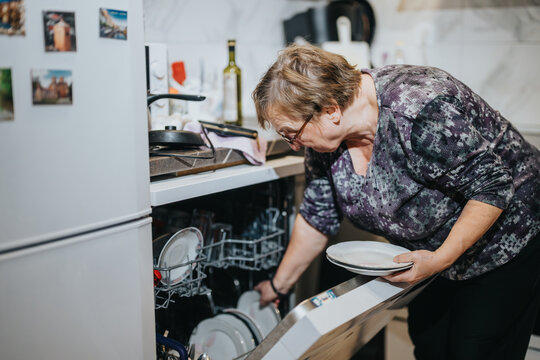 A senior woman actively engages in unloading a dishwasher, showcasing daily life, responsibility, and independence in a contemporary home environment.
