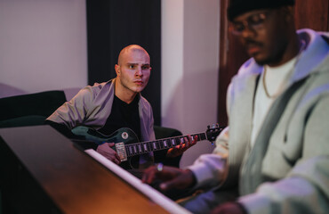 Two musicians collaborate with guitar and piano in a cozy studio setting. The focus is on...