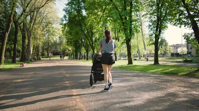 A young sporty woman walks in the park with her son in the running board. Mom is happy to do sports in the park with her newborn baby on a sunny day. Concept of power, fitness, family