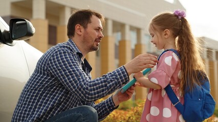 Smiling father giving textbook daughter pupil with backpack hugging before going school campus closeup. Happy dad embracing little female kid child arriving elementary education lesson at schoolyard