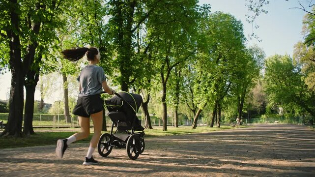 A young sporty woman runs in the park with her son in the running board. Mom is happy to do sports in the park with her newborn baby on a sunny day. Concept of power, fitness, family