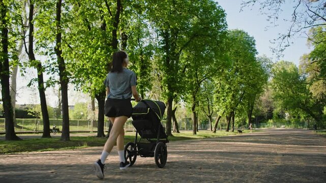 A young sporty woman runs in the park with her son in the running board. Mom is happy to do sports in the park with her newborn baby on a sunny day. Concept of power, fitness, family