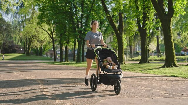 A young sporty woman runs in the park with her son in the running board. Mom is happy to do sports in the park with her newborn baby on a sunny day. Concept of power, fitness, family