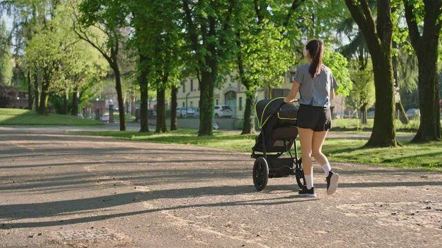 A young sporty woman walks in the park with her son in the running board. Mom is happy to do sports in the park with her newborn baby on a sunny day. Concept of power, fitness, family