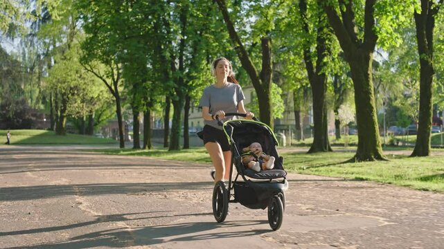 A young sporty woman runs in the park with her son in the running board. Mom is happy to do sports in the park with her newborn baby on a sunny day. Concept of power, fitness, family