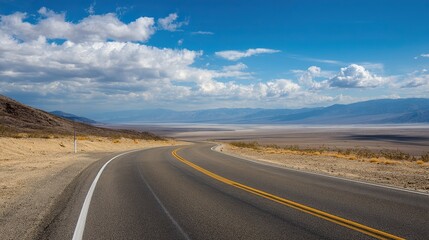 Fototapeta premium A long road winds through the desert with blue skies and white clouds. Ahead is the endless horizon and mountains in the distance, captured from behind with a wide-angle lens.
