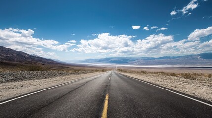 Naklejka premium A long road winds through the desert with blue skies and white clouds. Ahead is the endless horizon and mountains in the distance, captured from behind with a wide-angle lens.